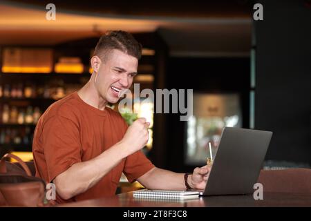 Aufgeregter junger Mann, der E-Mails mit guten Nachrichten liest, wenn er am Tisch im Restaurant sitzt Stockfoto