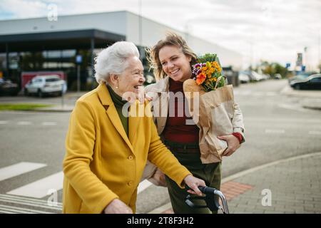 Reife Enkelin mit Großmutters Einkaufstasche. Seniorin und Betreuerin, die im kalten Herbst mit Lebensmitteln aus dem Supermarkt nach Hause gehen Stockfoto