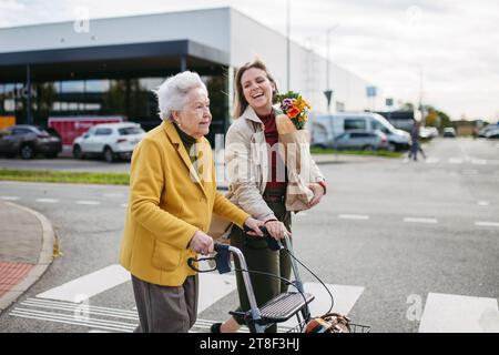 Reife Enkelin mit Großmutters Einkaufstasche. Seniorin und Betreuerin, die im kalten Herbst mit Lebensmitteln aus dem Supermarkt nach Hause gehen Stockfoto