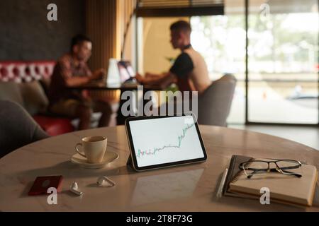 Börsendiagramm auf digitalem Tablet neben einer Tasse Kaffee und Planer auf dem Tisch, Händler arbeiten im Hintergrund Stockfoto