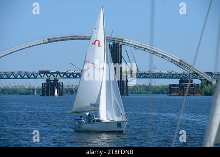 Segelyacht schwimmt auf dem Wasser des Flusses Dnipro, Brücke auf einem Hintergrund. August 2021. Kiew, Ukraine Stockfoto