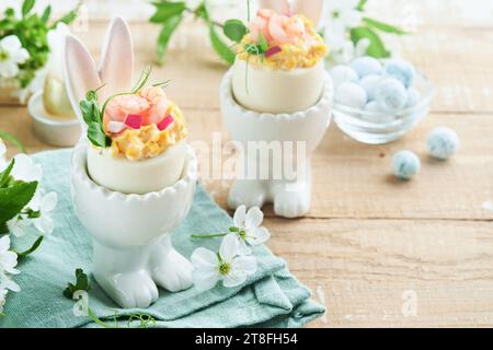 Gefüllte oder verteufelte Eier mit Eigelb, Garnelen, Erbsen-Mikrogrün mit Paprika in kaninchenförmigem Ständer für ostertisch schmücken frische Kirsch- oder Apfelblüten Stockfoto