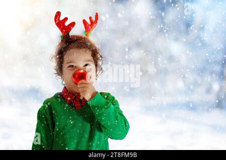 Kind mit Rentiernase und Geweih am Heiligabend. Ein kleiner Junge mit einer roten Kugel und einem Weihnachtsgeschenk. Kinder öffnen Weihnachtsgeschenke. Stockfoto