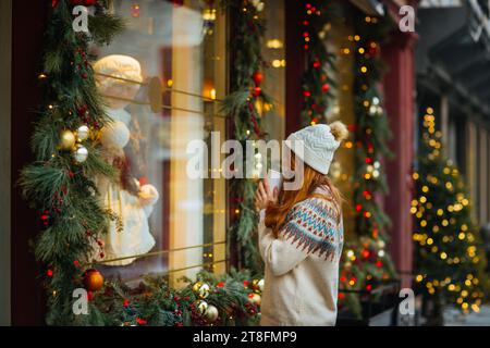 Eine Frau in einem warmen Pullover und einer Beanie genießt eine festliche Weihnachtsfensterung, während sie in Quebec, Kanada, eine Tasse hält Stockfoto