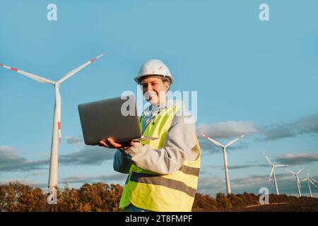 Eine Ingenieurin in Sicherheitsausrüstung verwendet einen Laptop mit Windturbinen im Hintergrund, die erneuerbare Energien und Technologie repräsentieren. Stockfoto