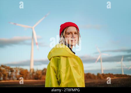 Eine fröhliche junge Frau, die in einer hellgelben Jacke und einer roten Mütze in die Kamera schaut, steht vor einem Windpark und symbolisiert nachhaltiges Wohnen und g Stockfoto