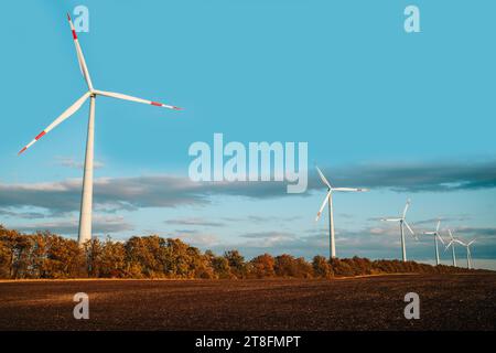 Eine ruhige Szene mit einer Reihe von Windturbinen vor einem leuchtend blauen Himmel mit herbstlichem Laub und einem frisch gepflügten Feld im Vordergrund. Stockfoto