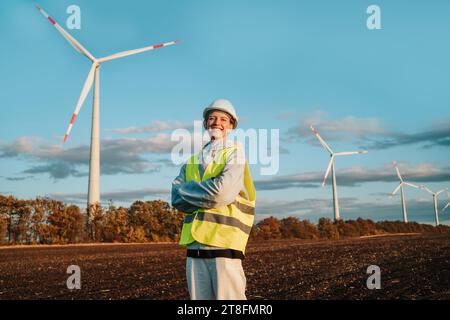 Eine freudige Ingenieurin mit gekreuzten Armen steht selbstbewusst auf einem Feld, Windturbinen und einem klaren blauen Himmel im Hintergrund, der erneuerbar ist Stockfoto