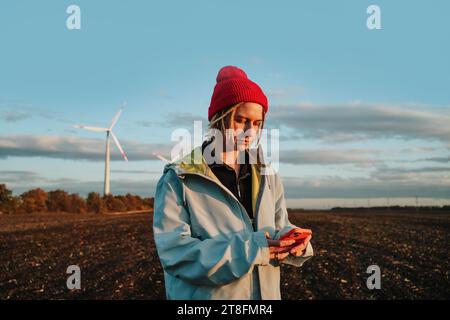 Eine alternative Frau, die ein Handy in einer Jeansjacke von Windturbines in einer Jeansjacke und einer roten Mütze verwendet, steht vor einer Windfarm und symbolisiert Sustai Stockfoto