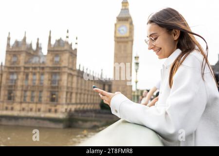 Eine junge lächelnde Frau in einem weißen Mantel benutzt ihr Smartphone vor dem legendären Big Ben und Westminster in London. Stockfoto