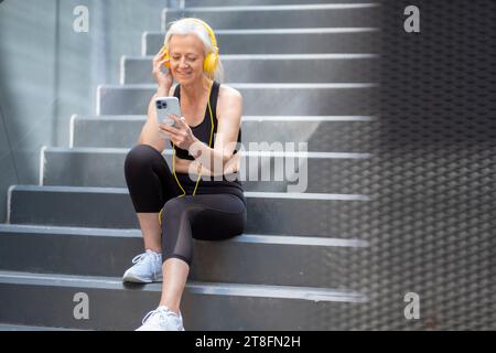 Eine lächelnde Seniorin sitzt auf einer städtischen Treppe, vertieft in ihr Smartphone mit gelben Kopfhörern in Trainingskleidung. Stockfoto