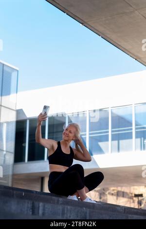 Eine fitte Frau in Sportbekleidung macht eine Pause, um mit ihrem Smartphone ein Selfie zu machen und sitzt auf einer städtischen Treppe unter klarem blauen Himmel. Stockfoto