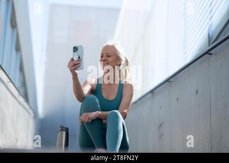 Eine glückliche Seniorin in Activewear macht ein Selfie mit ihrem Handy und ruht sich nach einer Fitness-Session im Freien aus. Stockfoto