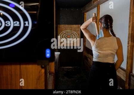 Eine junge Frau wird von hinten gefangen genommen, während sie sich darauf vorbereitet, eine Axt in einem schwach beleuchteten Innenbereich auf ein Ziel zu werfen, in dem Konzentration und Leis gezeigt werden Stockfoto