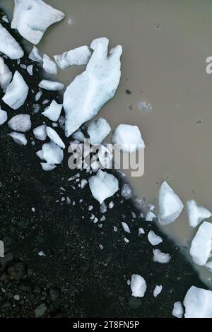 Aus der Vogelperspektive sind helle weiße Eisbrocken entlang des dunklen vulkanischen Sandes am Rande einer trüben Lagune im Vatnajokull-Nationalpark in Island verstreut Stockfoto