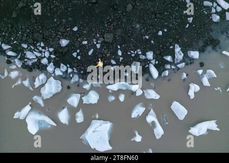 Von unten mit einer Person in gelber Jacke, die auf einem vulkanischen schwarzen Sandstrand steht, umgeben von verstreuten weißen Eisbrocken in Vatnajokull Stockfoto
