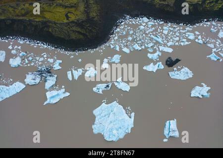 Luftaufnahme von zahlreichen weißen Eisstücken, die auf einem dunkelbraunen Gletscherfluss an der Küste schweben, im Kontrast zu rauem Gelände in Vatnajokull Nation Stockfoto