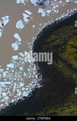 Von oben sehen Sie zahlreiche weiße Eisstücke, die auf einem dunkelbraunen Gletscherfluss an der Küste schweben, im Kontrast zu zerklüftetem Gelände in Vatnajokull Nationa Stockfoto