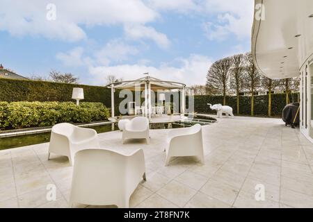 Weiße leere Stühle auf der Terrasse neben dem Swimmingpool und Efeu im Hinterhof des modernen Hauses vor bewölktem Himmel Stockfoto