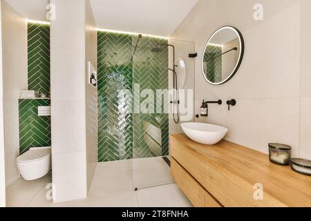 Duschkabine und Toilettenschüssel mit Waschbecken auf Holzschrank in stilvollem Badezimmer mit grünen und weißen Fliesen im modernen Haus Stockfoto