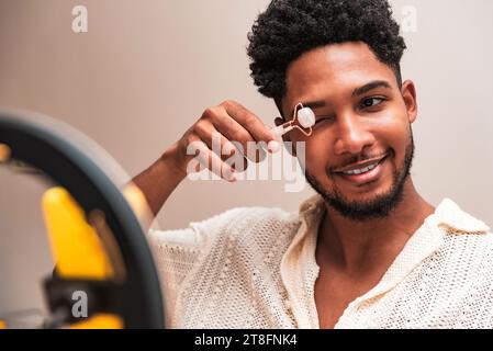 Ein junger lateinmann in einem lässigen Hemd benutzt einen Gesichtsroller, während er mit einem Ringlicht an einem Make-up-Tisch sitzt. Stockfoto