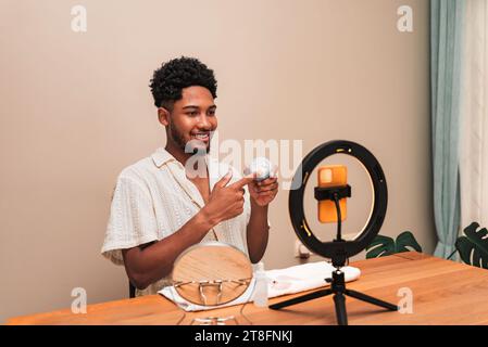 Ein fröhlicher junger Mann aus lateinamerika sitzt an einem Schreibtisch und nimmt sich mit einem Smartphone auf, das an einer Ringleuchte befestigt ist. Er hält ein Hautpflegeprodukt für eine Bewertung in der Hand. Stockfoto