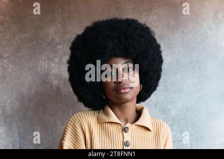 Porträt einer selbstbewussten jungen afroamerikanischen Frau mit lockigen dunklen Haaren, die die Kamera gegen eine verschwommene graue Wand anschaut Stockfoto