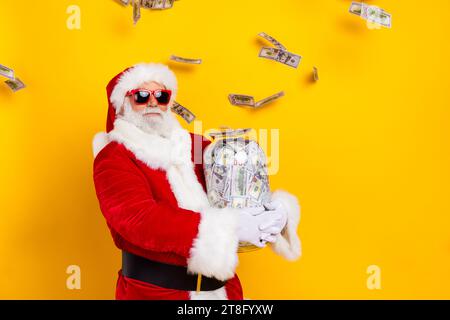 Foto des selbstbewussten weihnachtsmanns glückliche neue Ersparnisse mit großem Glasglas voller Banknoten Einnahmen isoliert auf gelbem Hintergrund Stockfoto