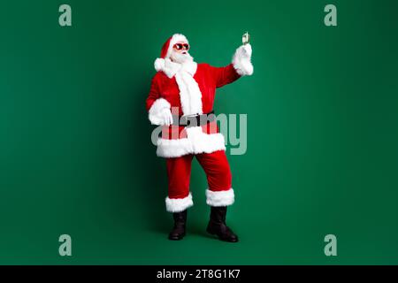 Foto in voller Länge von cool aufgeregten claus tragen rotes Kostüm sprechen weihnachtswünsche trinken Champagner isolierte grüne Farbe Hintergrund Stockfoto