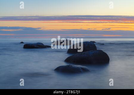 Romantische Atmosphäre bei friedlichem Sonnenuntergang am Meer. Große Felsbrocken, die aus glattem, welligem Meer ragen. Gelber Streifen am Horizont und winzige Wolken Stockfoto