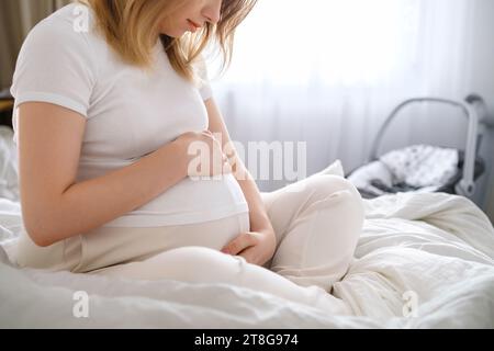 Schwangere Frau in leichten Kleidern sitzt auf dem Bett und umarmt sanft ihren Bauch. Stockfoto