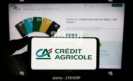 Person, die ein Mobiltelefon mit dem Logo des französischen Finanzdienstleisters Crédit Agricole SA vor der Webseite der Firma hält. Schwerpunkt auf dem Display des Telefons. Stockfoto