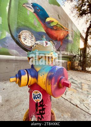 Miami, Florida, USA. Januar 2023. Farbenfroher, mit Graffiti bedeckter Feuerhydrant auf der Straße neben einem Vogelgemälde. Wynwood ist die Heimat der höchsten Konzentration an Graffiti Street Art in den USA. Wandmalereien aus aller Welt zeigen ihr Talent an Wänden mit überlebensgroßen Meisterwerken. Wynwood ist bekannt für seine vielen farbenfrohen Wandmalereien und eines der beliebtesten Viertel der Stadt. Das Wynwood Walls ist ein Freiluftmuseum, in dem große Werke einiger der weltweit bekanntesten Straßenkünstler ausgestellt werden. In den umliegenden Straßen wurden Lagerhäuser mit Handwerksbrauereien und ausgefallenen Kunstgalerien umgebaut. Hallo Stockfoto