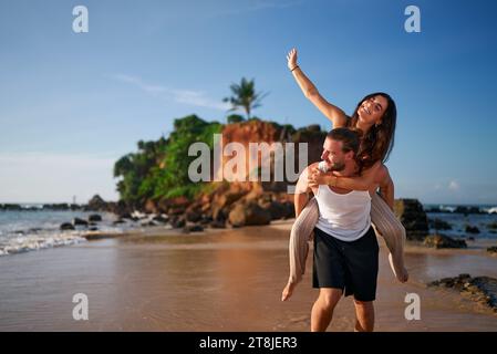 Fröhliches Paar genießt einen Huckepack-Ritt am tropischen Strand, die Frau hebt die Hand hoch, der Mann trägt den Partner auf dem Meer. Romantischer Kurzurlaub, verspielte Stimmung, Sandstrand Stockfoto