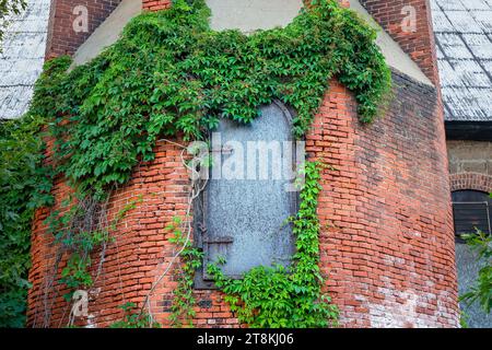 Eine eiserne Zugangstür und grüner Efeu am Fuße eines alten industriellen Ziegelschornsteins. Viel Textur. Die Konzepte könnten die Vintage-Industrie, Architec umfassen Stockfoto