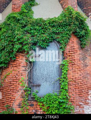 Eine eiserne Zugangstür und grüner Efeu am Fuße eines alten industriellen Ziegelschornsteins. Viel Textur. Die Konzepte könnten die Vintage-Industrie, Architec umfassen Stockfoto