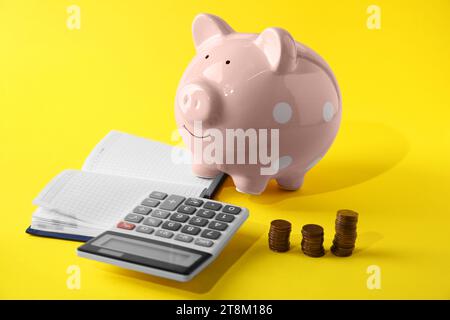 Taschenrechner, rosafarbene Schweinebank, Notizbuch und gestapelte Münzen auf gelbem Hintergrund Stockfoto