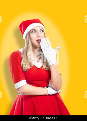 weihnachtsmann mit überraschtem Ausdruck auf gelbem Hintergrund. Junge lateinblonde Frau im roten santa-Anzug, die ihre Hand an den Mund legt Stockfoto