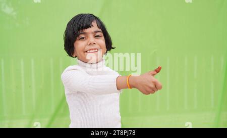 Glückliches positives kleines Mädchen in weißem Rollkragen, das Daumen nach oben wie Geste zeigt, isoliert auf einfachem Pastellhintergrund hellgrünem Studioporträt. Stockfoto