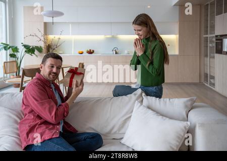 Interessierter Mann erhält Geschenkbox von liebevoller Frau, die auf der Couch in der Nähe mit gefalteten Palmen wartet Stockfoto