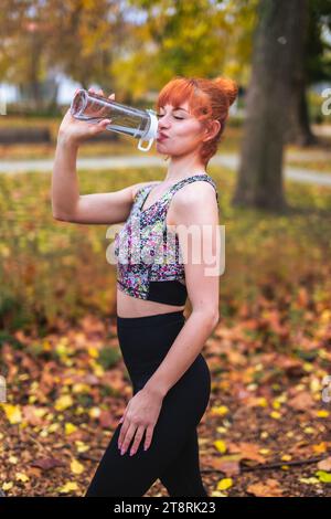 Rothaarige 20er Jahre Kaukasierin trinkt aus der Flasche, nachdem sie im Spätherbst im Park trainiert hat Stockfoto