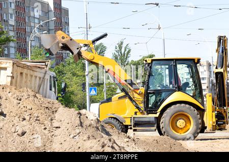 Der verschiebbare Eimer eines Straßenbaggers lädt an einem Sommertag Sand in die Rückseite eines Lkws vor dem Hintergrund einer Stadtstraße. Stockfoto