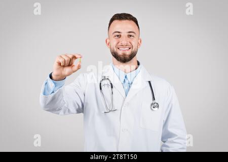 Ein lächelnder junger Arzt zeigt Pille, weißes Fell, grauer Hintergrund Stockfoto