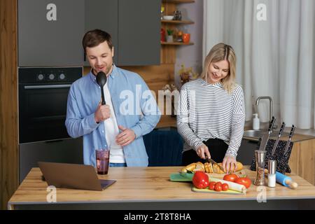 Heimkonzert. Verspielte Ehepartner, die Spaß in der Küche haben, singen und tanzen. Fröhliches Paar, das Küchenutensilien als Mikrofone benutzt und beim Kochen täuscht Stockfoto