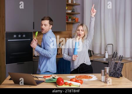 Heimkonzert. Verspielte Ehepartner, die Spaß in der Küche haben, singen und tanzen. Fröhliches Paar, das Küchenutensilien als Mikrofone benutzt und beim Kochen täuscht Stockfoto