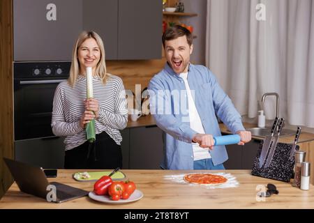 Glücklicher junger Mann und Frau lachen und singen mit Küchengeräten, die zu Hause zusammen kochen. Ein überfreuliches lächelndes Paar hat Spaß beim Zubereiten des Essens Stockfoto