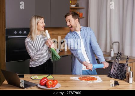 Glücklicher junger Mann und Frau lachen und singen mit Küchengeräten, die zu Hause zusammen kochen. Ein überfreuliches lächelndes Paar hat Spaß beim Zubereiten des Essens Stockfoto