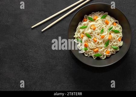 Instant-Nudeln, gekocht mit Gemüse, in einer grauen Schüssel und Essstäbchen, auf dunklem Hintergrund, Draufsicht, Platz zum Kopieren von Text. Stockfoto