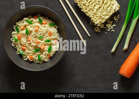 Instant-Nudeln gekocht und roh, mit grünen Zwiebeln, Karotten und Stäbchen, auf dunklem Hintergrund, Draufsicht, Platz zum Kopieren von Text. Stockfoto