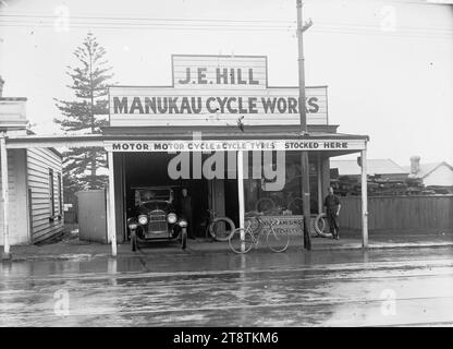 J E Hill, Manukau Cycle Works, Onehunga, View of Manukau Cycle Works, Victoria Street, Onehunga (Eigentümer J E Hill) mit einem Auto, das im Eingang der Garage auf der linken Seite des Ladens geparkt ist. Ein Fahrrad lehnt sich an die Verandastütze vor dem Laden, und ein Mann steht rechts und blickt auf den Fotografen. Die Werbung auf der Ladenfront lautet "Motor-, Motorrad- und Fahrradreifen hier vorrätig" und "Vulkanisieren einer Spezialität". Eine Norfolk-Kiefer wächst hinter dem nebenan gelegenen Grundstück. Hinter dem Zaun des Nachbargrundstücks ist Holz gestapelt Stockfoto